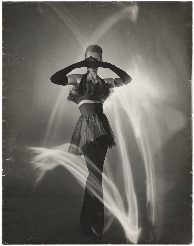 a black and white phtograph of supermodel Lisa dress in a coupe tutu, covering her eyes with her hands, which are dressed in shoulder length black satin gloves. a wisp of white light dances around her