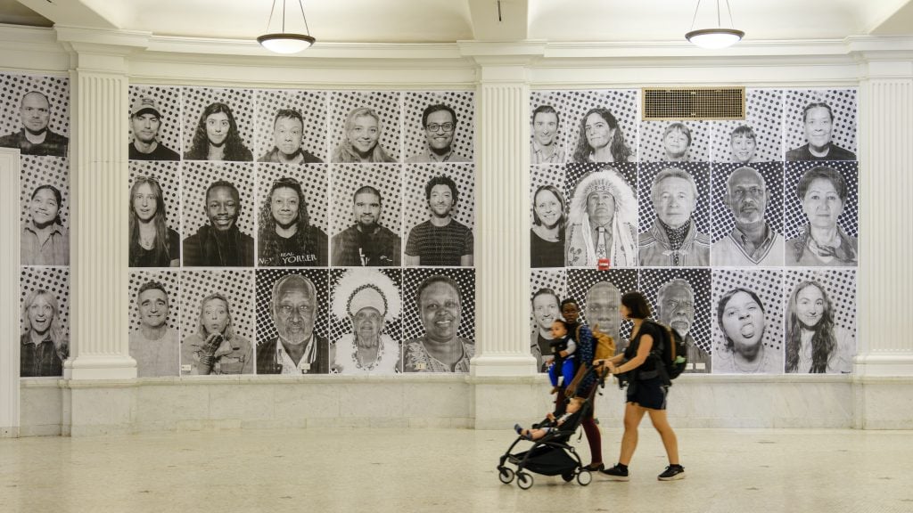 An installation of portraits taken by JR's "Inside Out Project" photo booth truck for "Portraits on Climate and Health: Dreams We Carry" on view in the Ellen V. Futter Gallery at the American Museum of Natural History in New York. The black and white portraits of a group of people of various ages and races, some of which are of Indigneous people in traditional garb, is installed in a white-walled museum gallery on either side of a pilaster, with two moms walking by with their babies, one in a stroller, the other strapped to her chest.