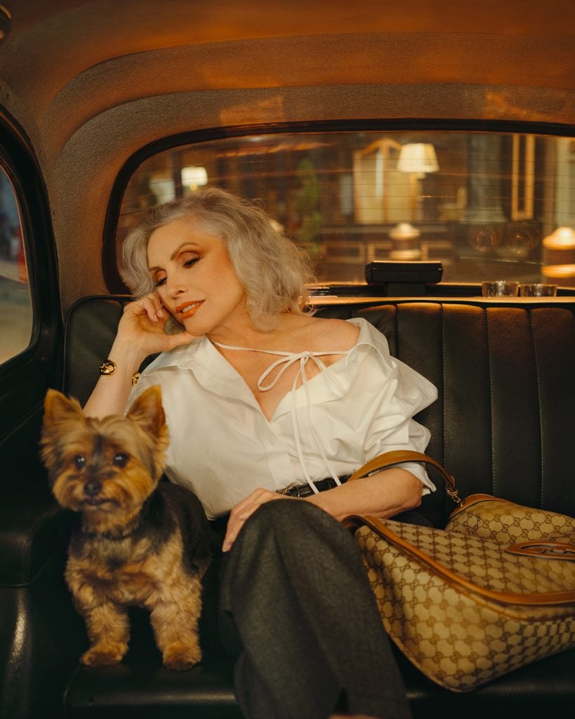 a woman in a white blouse sits in the back of a taxi with a small dog