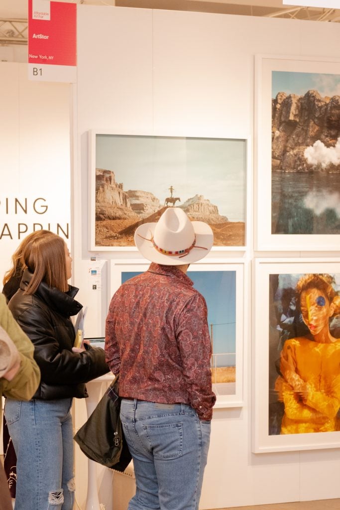 Two visitors to Affordable Art Fair NYC observe a series of artworks on a gallery booth wall.