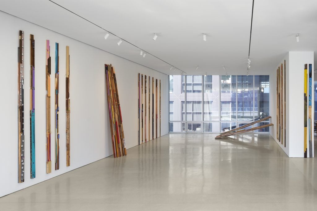 A white gallery space with work by Lubaina Himid installed, comprised of vertical colorfully painted sticks installed upright on the walls; the back wall of the gallery space is a wall of windows looking out to the building across the road.