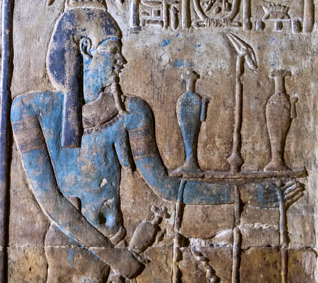 A photograph of a blue-tingedcarving of a figure standing before a table with vases on it as seen on an ancient wall of the Temple of Edfu.