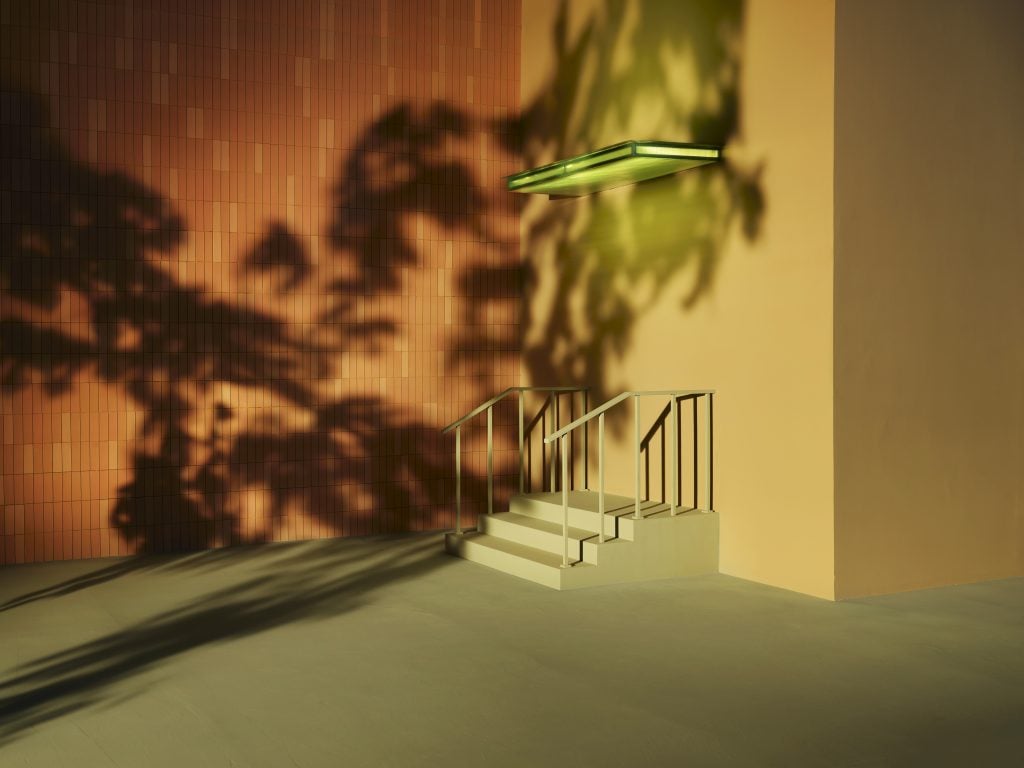 Photo of a staircase attached to a wall, with no entrance. The wall is beige and a shadow of a tree is cast on the wall.