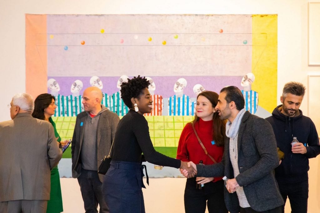 Artists and museum goers greet each other and talk in front of a large artwork on the wall