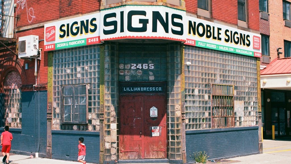 An old-fashioned looking storefront with glass block windows and a red door. The sign is for Noble Signs.