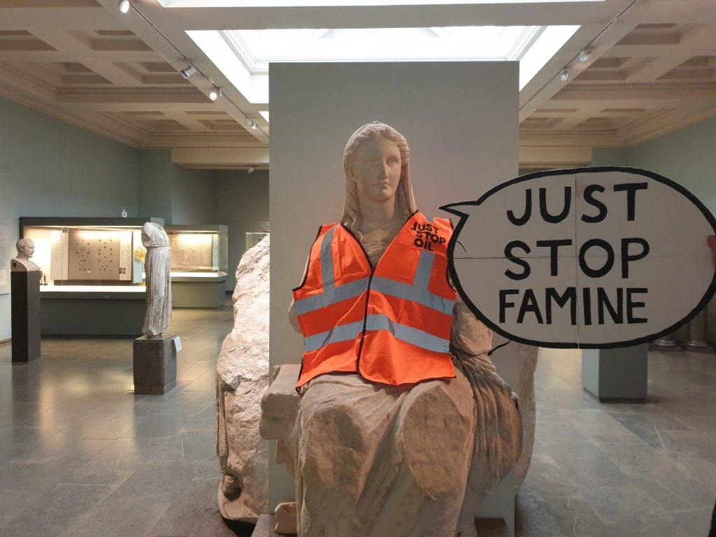 interior of a museum shows a seated ancient marble statue of a woman that is draped in an orange high-vis jacket, next to her is a speech bubble reading 'just stop famine'