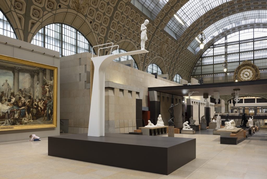  A contemporary white statue of a boy looking at his smartphone stands in the Musée d’Orsay’s grand sculpture hall. Surrounding the statue are classical bronze and marble sculptures, creating a contrast between old and new interpretations of the human form.