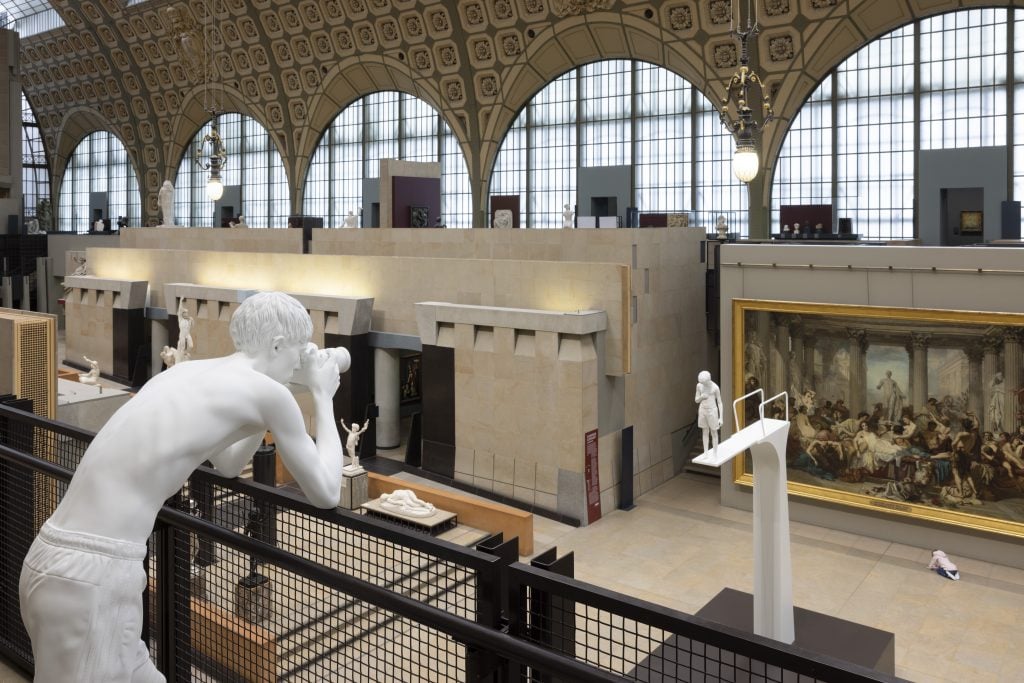 A white statue of a boy, peering through binoculars, overlooks the gallery of the Musée d’Orsay. Below, other contemporary sculptures are installed alongside classical marble works and a large neoclassical painting in the museum’s grand hall.