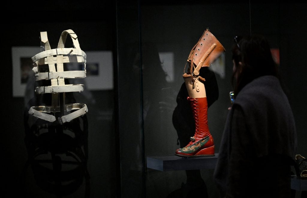 a display cabinet in a darkened space, showing a metal brace to be worn around a human torso and a prosthetic leg that appears to be wearing a red boot