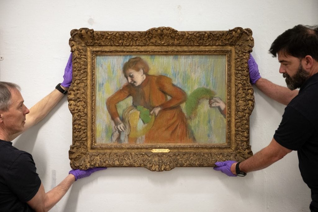 Two people wearing purple gloves carefully hang an ornate framed painting by Edgar Degas, depicting a woman in a red dress.