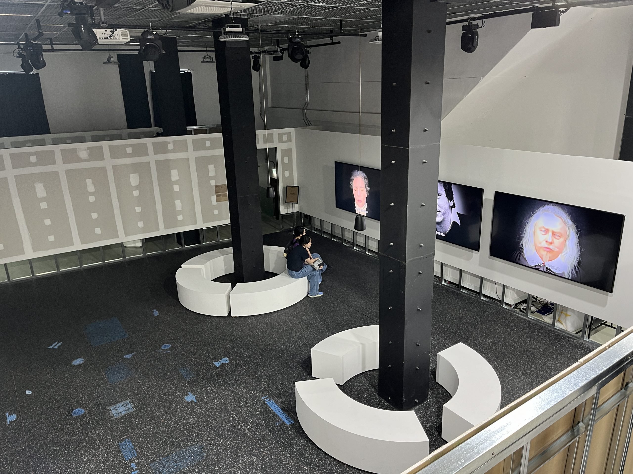 Three video screens hang on a white wall in a cavernous space with columns.