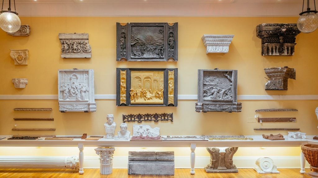 The Cast Hall at the Institute of Classical Architecture and Art. An array of plaster casts of columns and classical relief sculptures are displayed in a yellow room.