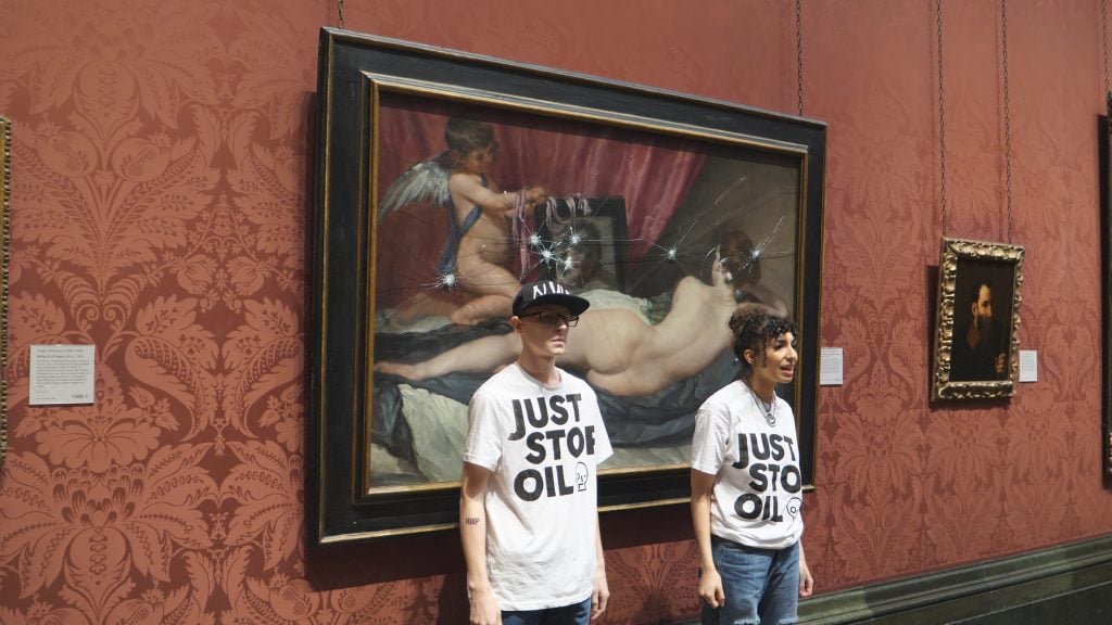 Two man in white T-shirts standing in front of a large painting of a naked woman