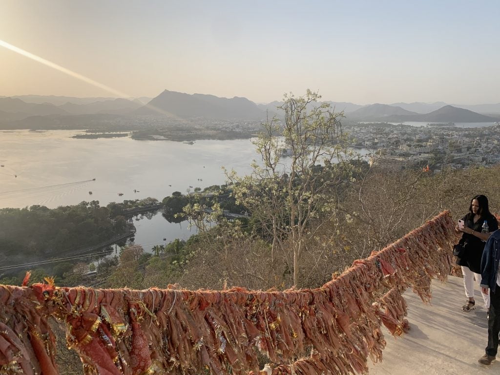 A beautiful landscape photograph of Udaipur, taken from atop a hill with a large body of water and hills in the distance below. There is a bannister decorated with tattered strips of orange fabric on the stairs in the foreground.