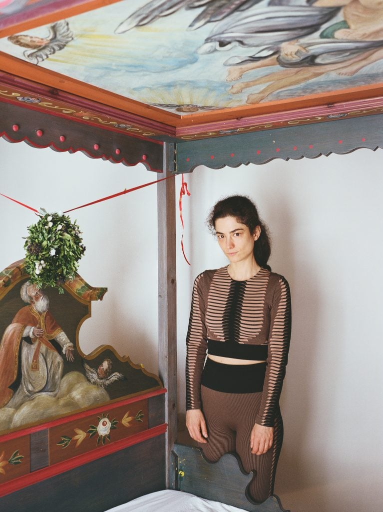 Portrait of Agnes Scherer standing behind an elaborately painted wooden canopy bed frame wearing a coordinated athleisure set.