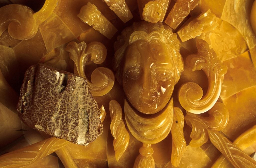 A rough piece of amber sits on top of a carved work of amber showing a boy's face amid flourishes