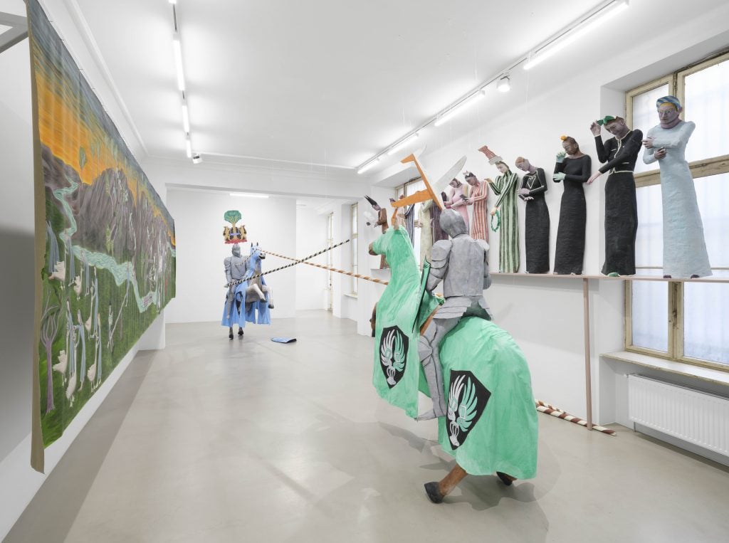 Installation view of Agnes Scherer's exhibition Savoir Vivre at ChertLüdde showing a row of human-scale medieval ladies along the wall of the gallery space and in the center two horses with fully armored jousters.