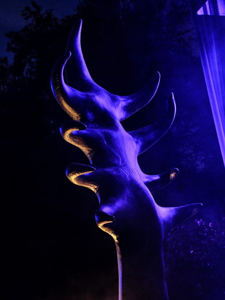 a large statue of an undefined shape with multiple tendrils reaching out placed on a pedastal in a dark room with neon purple lighting