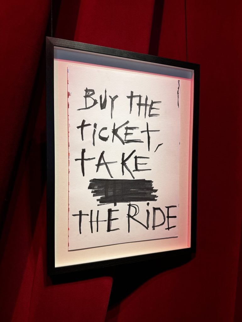 Artwork by Johnny Depp: a white paper scrawled with the words "Buy the ticket, take the ride" in a black frame hanging against a red curtain