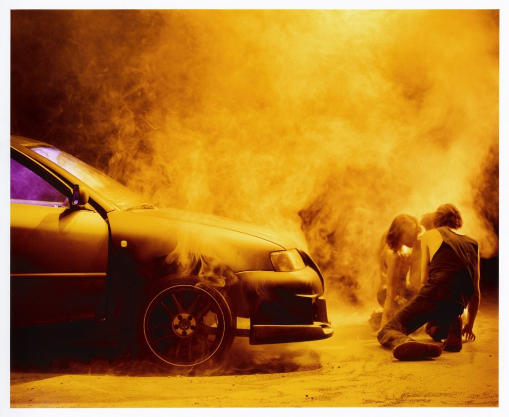 An image included at the forthcoming Paris Photo by Spector Books of the front half of a sedan car in front of which is two figures crouched down, and everything is surrounded in orangey-yellow spoke.