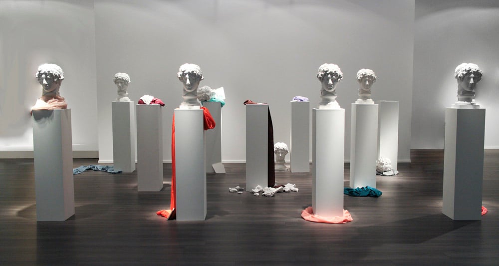 Installation view of Arte Povera artist Giulio Paolini's work comprised of a few rows of white pedestals with half having a marble bust on top, and various colorful scarfs draped on and around works.