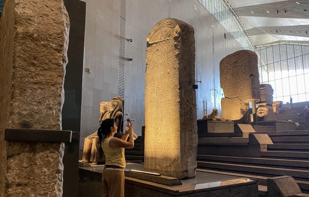 A woman holding up her smart phone at a large monument in a grand museum hall