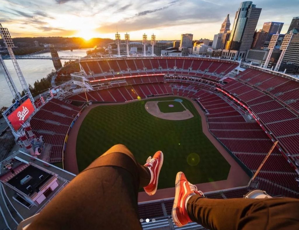 Isaac Wright's photograph of Great American Ballpark in Cincinnati. The artist photographed his legs dangling over the edge of the stadium, with the field below him and the city skyline beyond, sun rising over the horizon and the river.