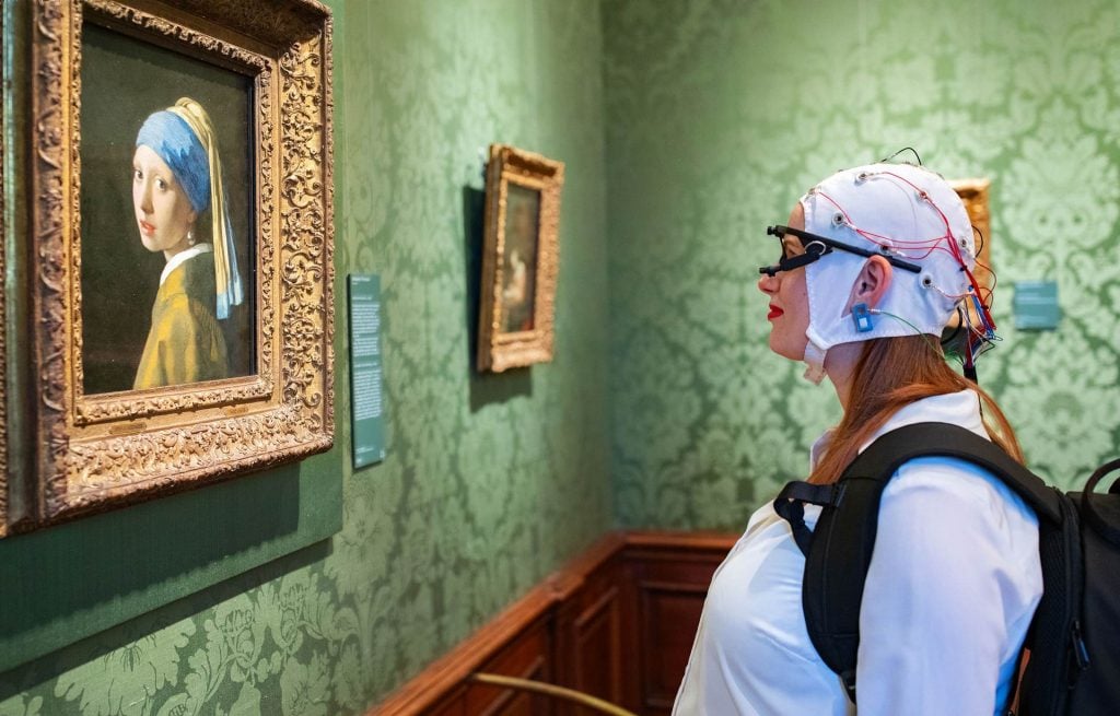 A woman wearing eye trackers and an EEG headset looking at Vermeer's painting Girl With Pearl Earrning