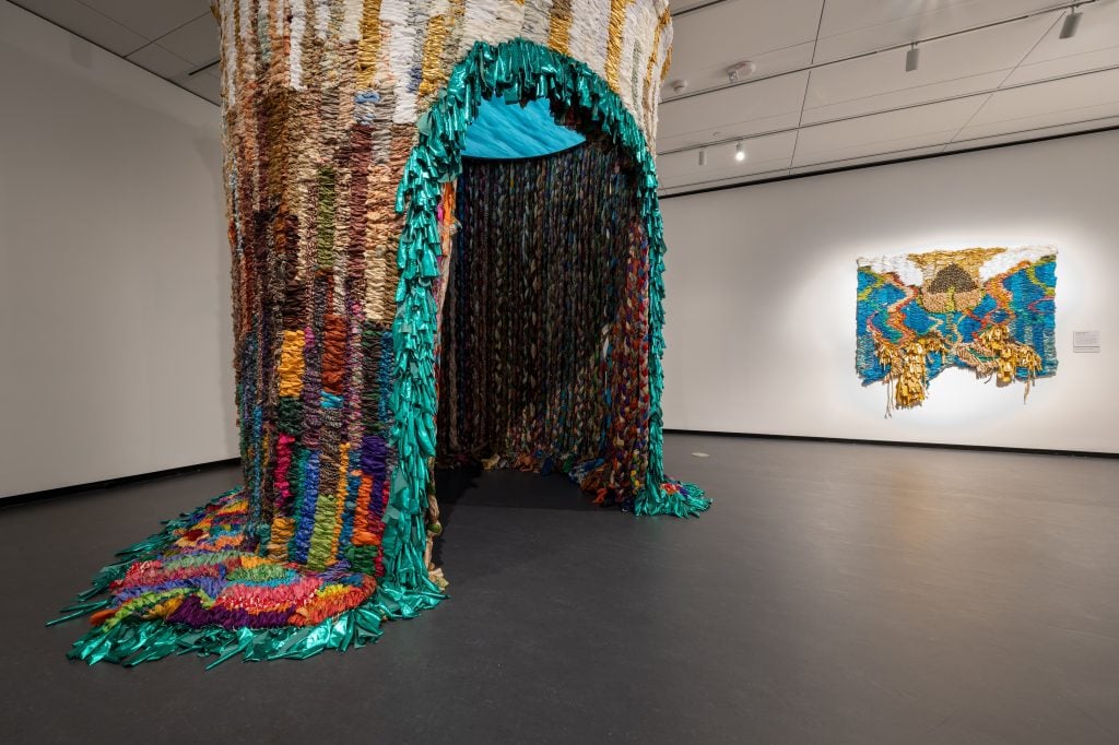 Installation view of “Suchitra Mattai: Myth From Matter” at the National Museum of Women in the Arts, Washington, D.C., showing her large-scale sculpture Siren Song, a round structure hanging from the ceiling with an arched entryway. It is made from long woven and braided strips of fabric taken from colorful saris. Another artwork hangs on the gallery wall.