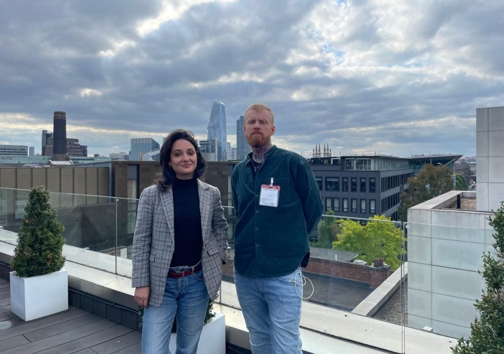 two people stand on a rooftop against an urban backdrop and a cloudy, dark grey sky