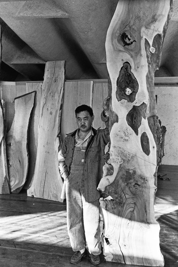 a black and white photo of a man in overalls standing in a studio next to a giant slab of standing wood