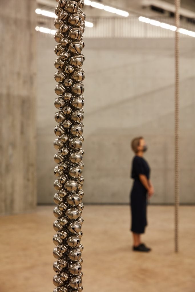 in the foreground, a rope made of bells hangs from the ceiling of a large gallery space. a woman can be seen standing in the background 