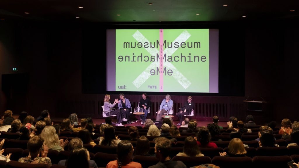 a large dark room with rows of seating in which we can see a sizable audience from behind, on the stage are five people set up like a discussion panel and over their heads is a projection reading 'Museum Machine Me' overlayed with a big 'X'