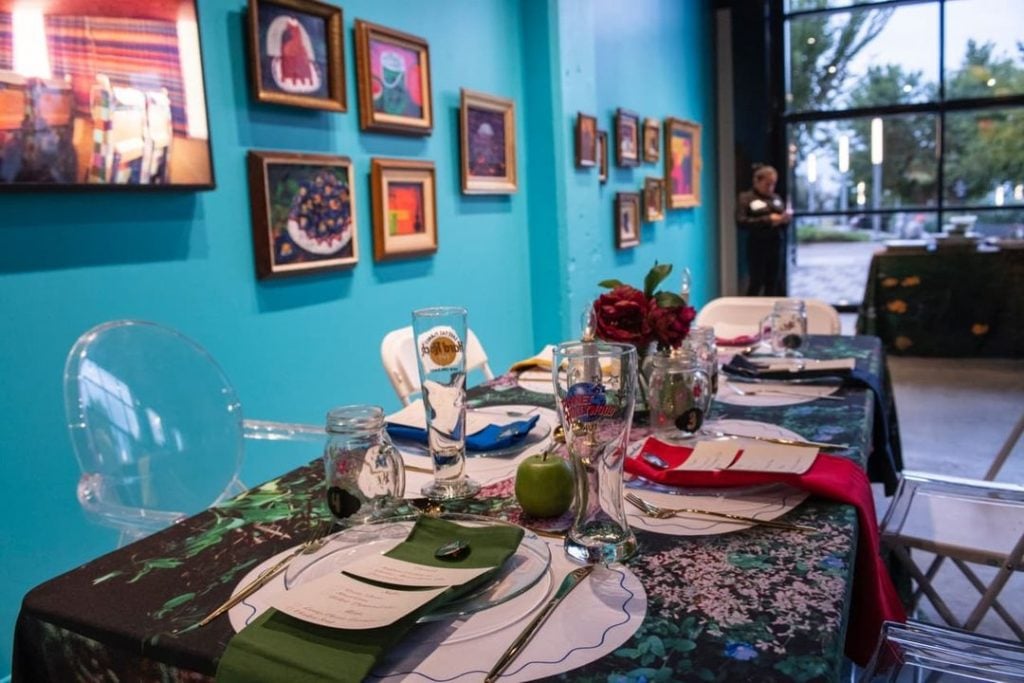 Azikiwe Mohammed designed the tablescapes for the opening reception for his show at Project for Empty Space Ironside. The tablecloth features a photograph by the artist, and there are many plates and glasses and flower arrangements on the table, with the artist's framed still life paintings hung on the robin's egg blue wall in gold frames. 