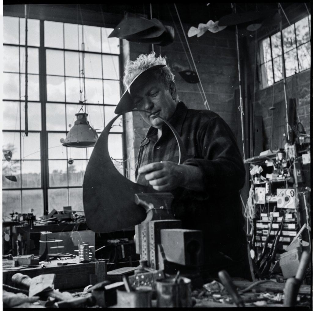 black and white photo of an elderly bearded man in a shirt working with tools on some kind of sculpture inside his messy studio