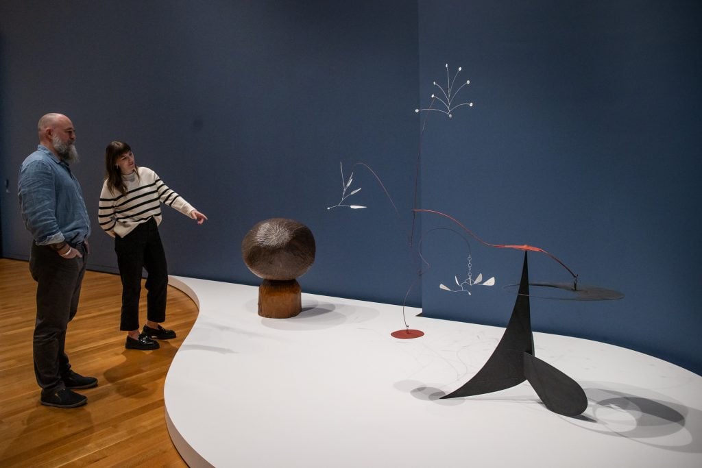 two people walking through a museum looking at as group of small statues of varying shapes displayed on a white table cloth in front of a dark blue wall