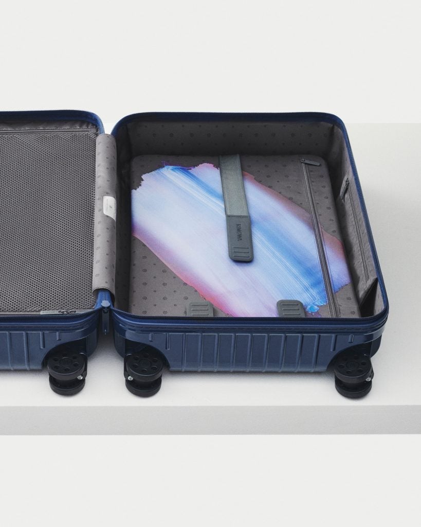 A photograph of a deep blue suitcase laying on the floor opened to review an artwork consisting of a single blue and lavendar colored brushstroke inside.