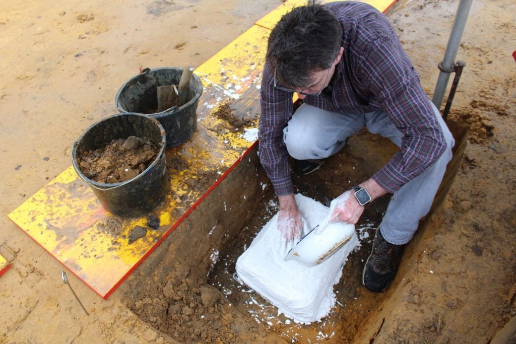 A photograph of an archaeologist applying plaster to a block of earth in a small dirt pit.