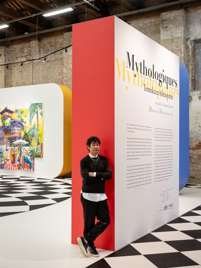 Artist Tomokazu Matuyama within his solo show "Mythologiques" leaning on one of the floating walls with the edge painted in red, and along the flat side the show title and didactic information.