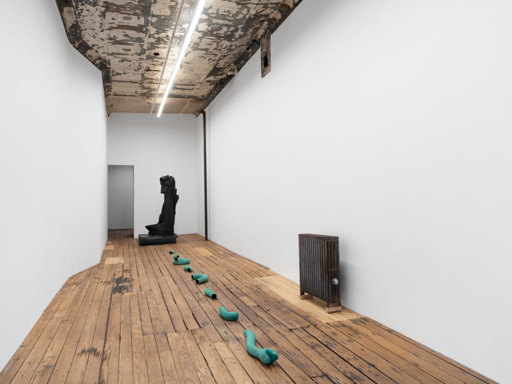 a gallery has turquoise metal sculptures on the uneven wood floor and an illunnaited neon light and an exposed radiator 