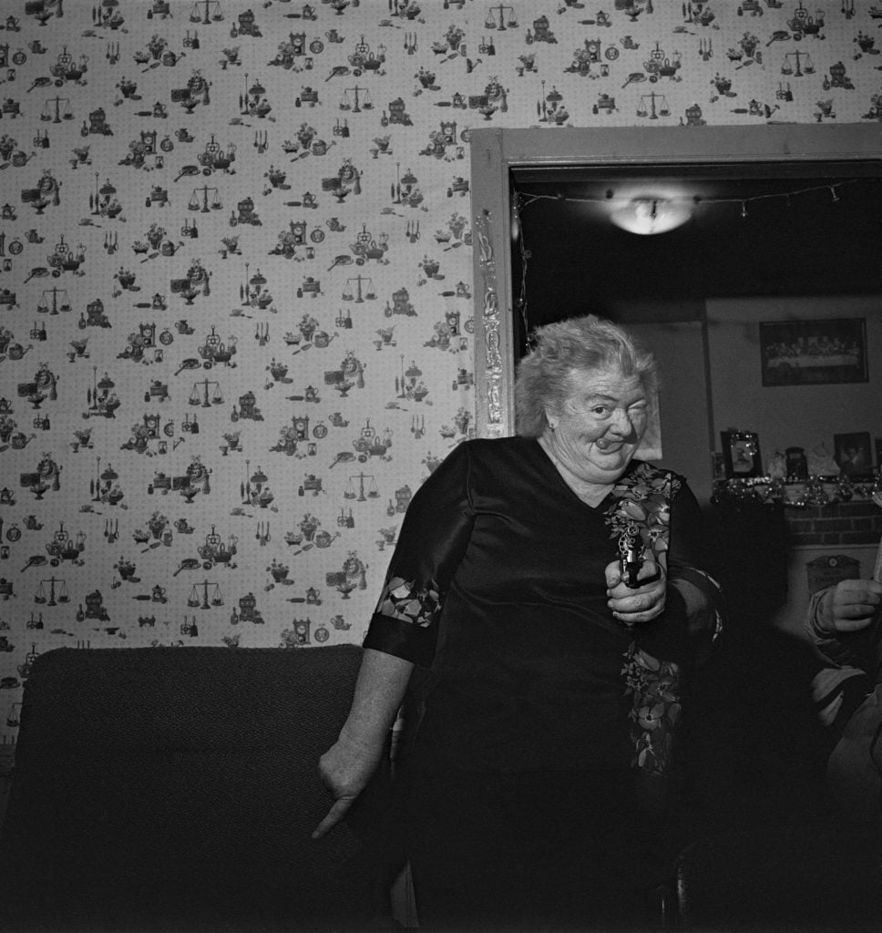 A black-and-white photo of an older woman in a dark outfit, smiling and pointing playfully with one hand while holding a small object in the other. She stands in front of a patterned wallpaper and a doorway, with part of a chair visible in the foreground. The scene is lively, suggesting a candid moment in a domestic setting.