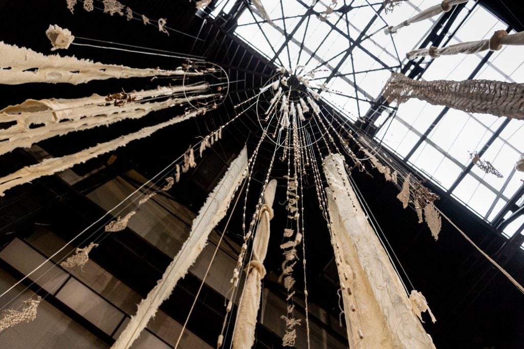 A photo of an artwork seen from below, hanging from a high ceiling, with various materials woven together