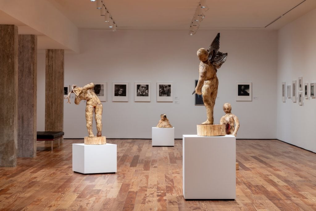 A photograph of an art exhibition room featuring sculptures and framed photographs. The sculptures, displayed on white pedestals, depict human-like figures with raw, textured surfaces, including a winged figure and another holding antlers. Framed black-and-white portraits hang on the walls in the background. The gallery has a minimalist design with wooden floors, concrete columns, and soft, focused lighting.