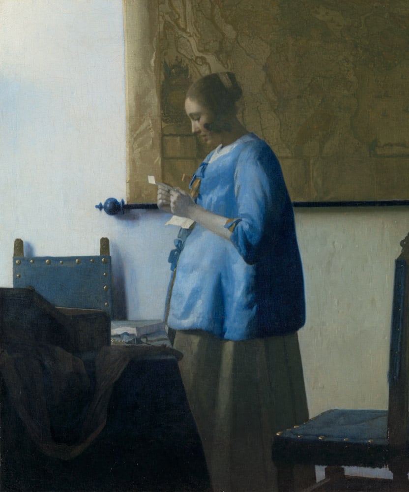 A serene woman, dressed in blue, intently reads a letter near a window.