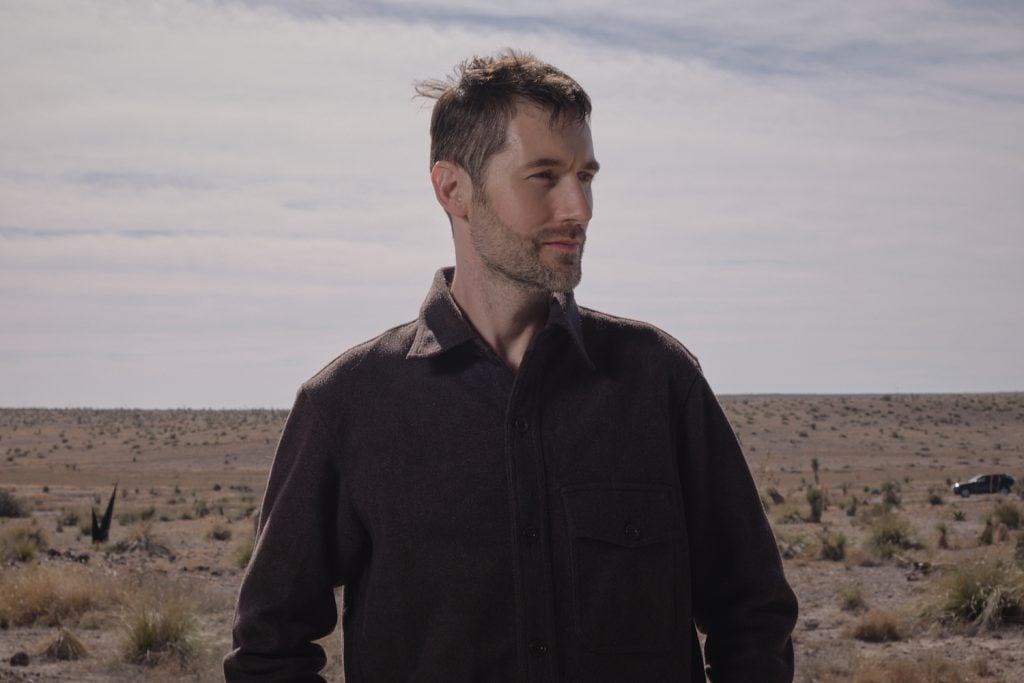 a man in a black shirt stands in the desert in a portrait