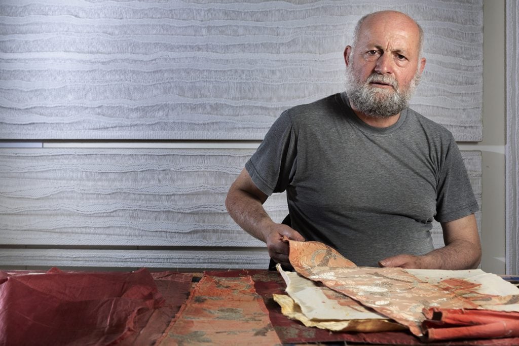 A caucasian man, bald, in grey beard, in a grey-green tee shirt, sitting at a wooden desk, with a stack of ancient fabrics in front of him.