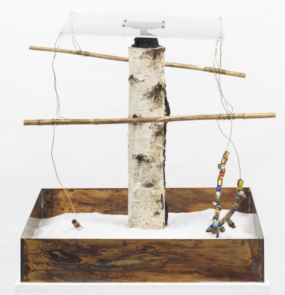 a small makeshift style contraption with a central pillar that rotates, dragging extensions from which hang wires and beads that make circular patterns in the sand base