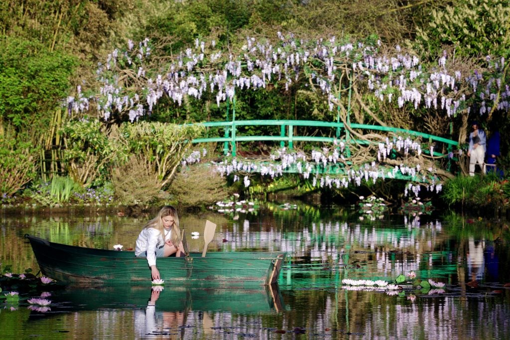 Camille Razat as Camille visiting Claude Monet's gardens at Giverny in Emily in Paris. A blonde woman in a white top siting in a green row boat reaches down toward a light pink water lily in a glassy pond in which reflections of the surrounding flowers and foliage are visible. The green Japanese bridge can be seen behind her.