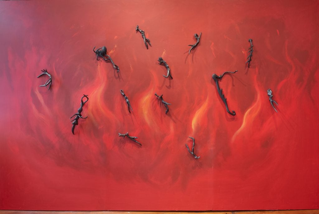 A series of small, vaguely figurative Helen Evans Ramsaran bronzes hanging on a wall painted red with orange flames.