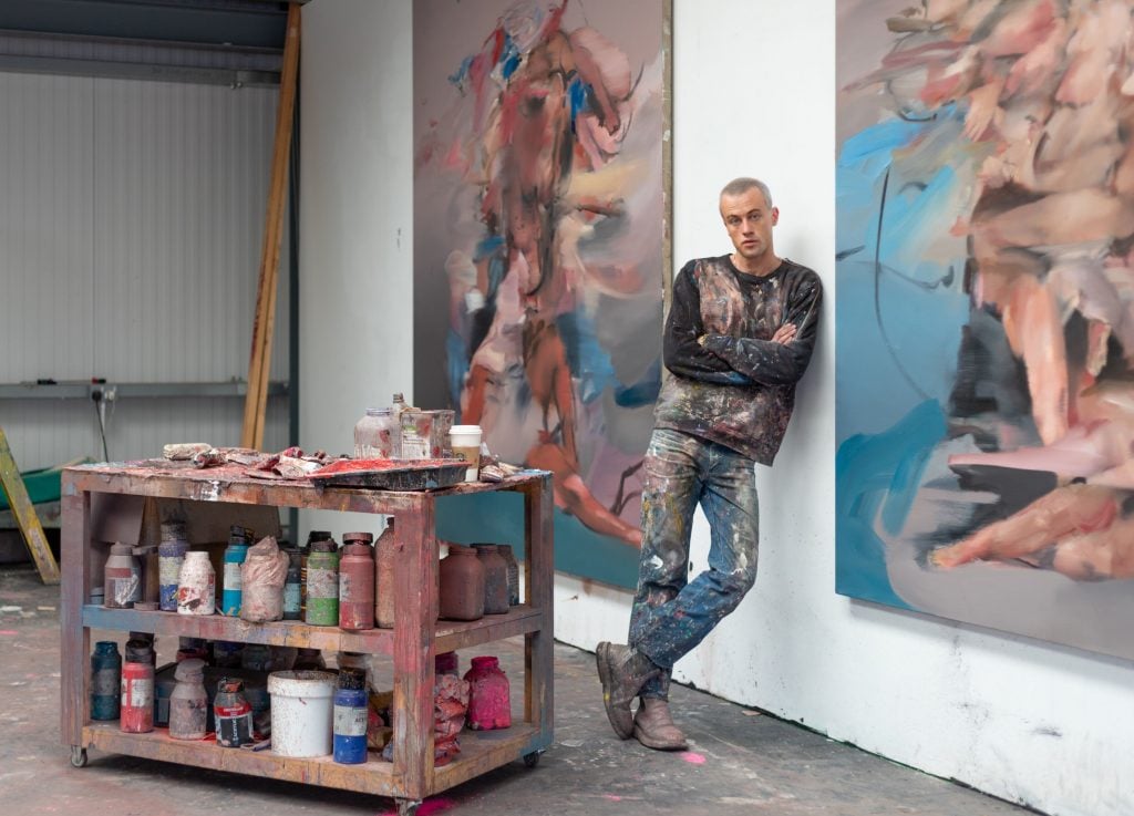 a man wearing blue jeans and a black jumper splattered in paint leans against the wall of an interior space, positioned between two large paintings that he is the author of. In the middle of the eroom is a moveable wooden tray stacked with bottles of paint and other painting materials